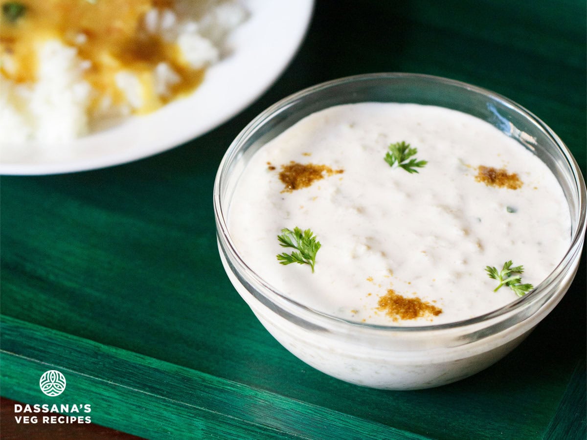 lauki ka raita served in a glass bowl sprinkled with roasted cumin powder and coriander leaves on a dark green tray.