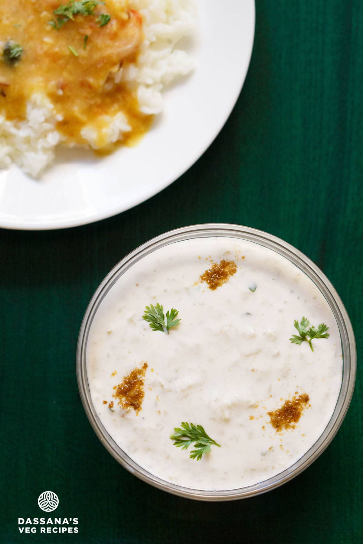 lauki ka raita in a glass bowl sprinkled with roasted cumin powder and coriander leaves on a dark green tray with a plate of dal and rice on top left side.