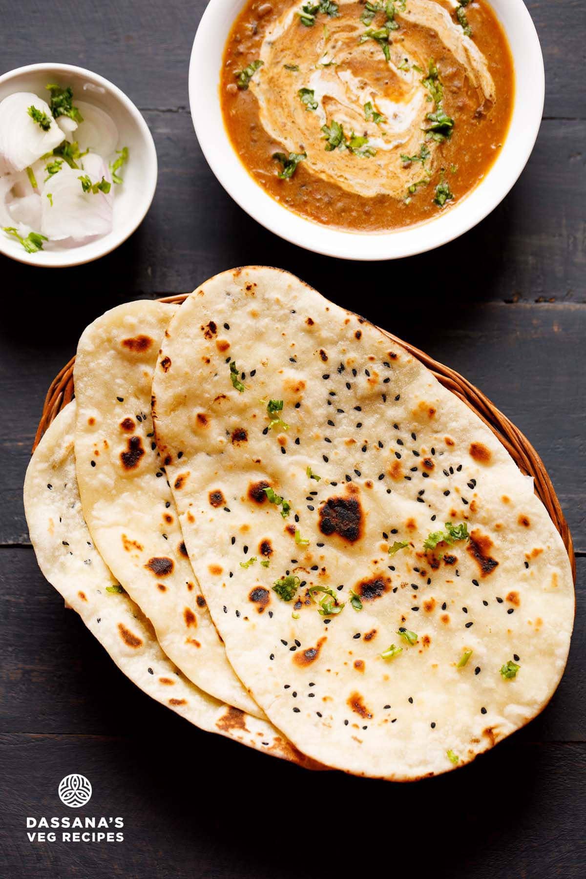 naan kept on top of each other in a cane basket. served with dal makhani in a white bowl and side of sliced onions garnished with coriander leaves in a small bowl.