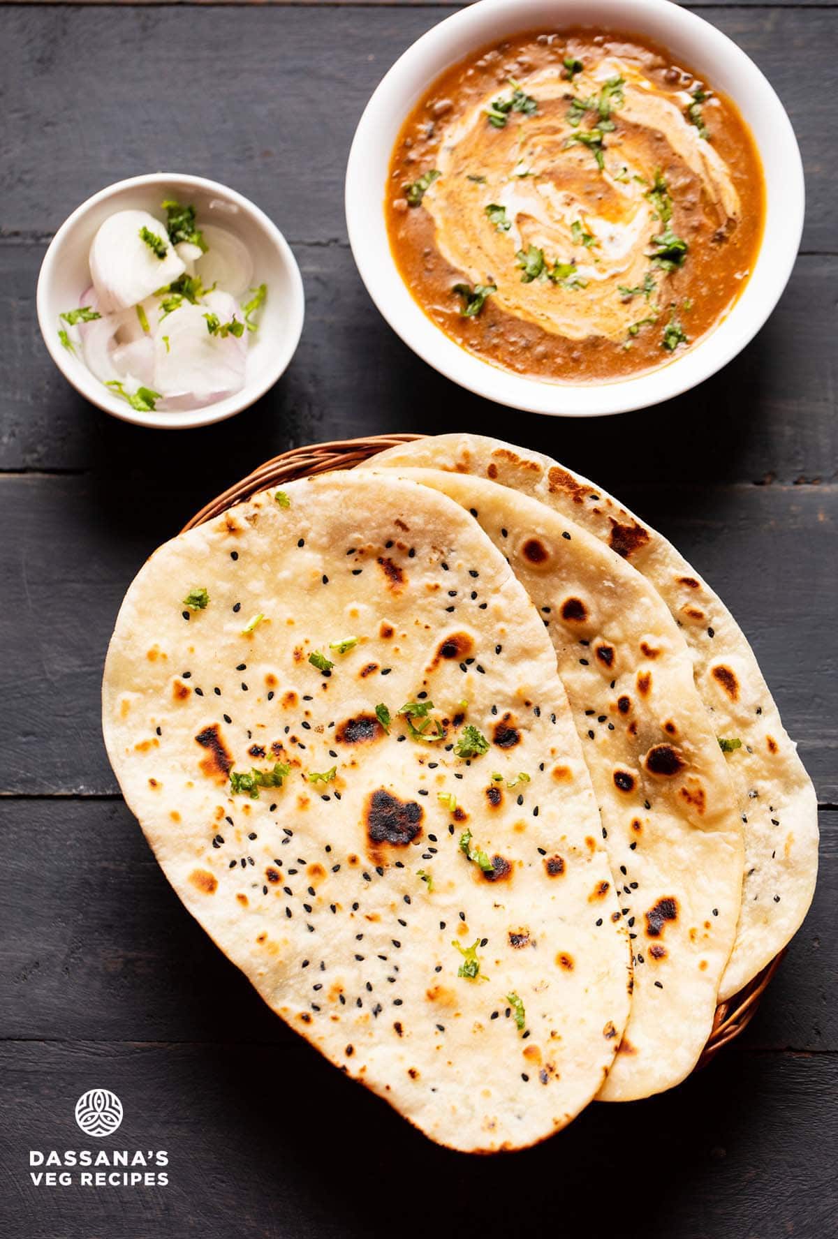 naan bread kept on top of each other in a cane basket. served with dal makhani in a white bowl and side of sliced onions garnished with coriander leaves in a small bowl.
