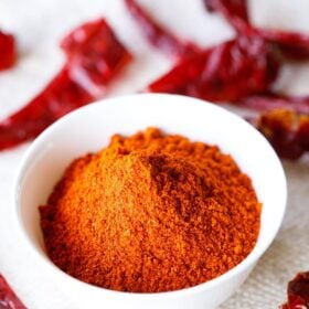 red chilli powder in a white bowl with dried red chillies on top and at the bottom of the bowl on a jute cloth.