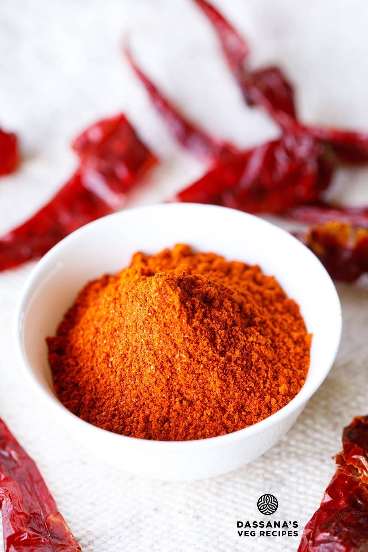 red chilli powder in a white bowl with dried red chillies on top and at the bottom of the bowl on a jute cloth.