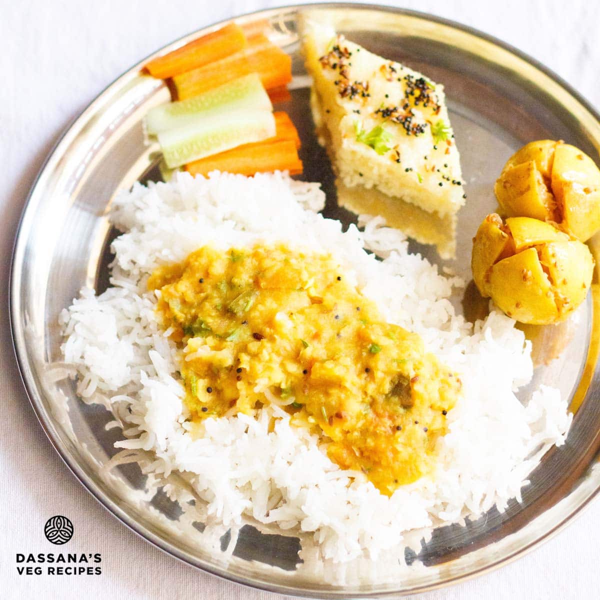a thali with tomato dal poured on steamed rice, lemon pickles, rava dhokla, carrot and cucumber sticks.