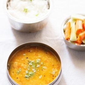 tomato pappu in a steel bowl with another bowl of steamed rice on top and a third bowl of carrot and cucumber sticks on top right side on a white napkin.