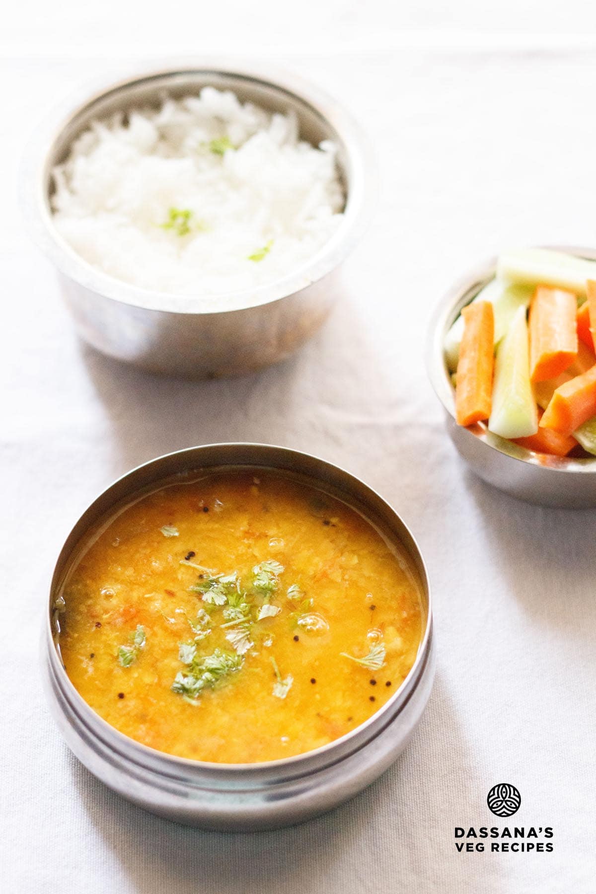 tomato pappu in a steel bowl with another bowl of steamed rice on top and a third bowl of carrot and cucumber sticks on top right side on a white napkin.