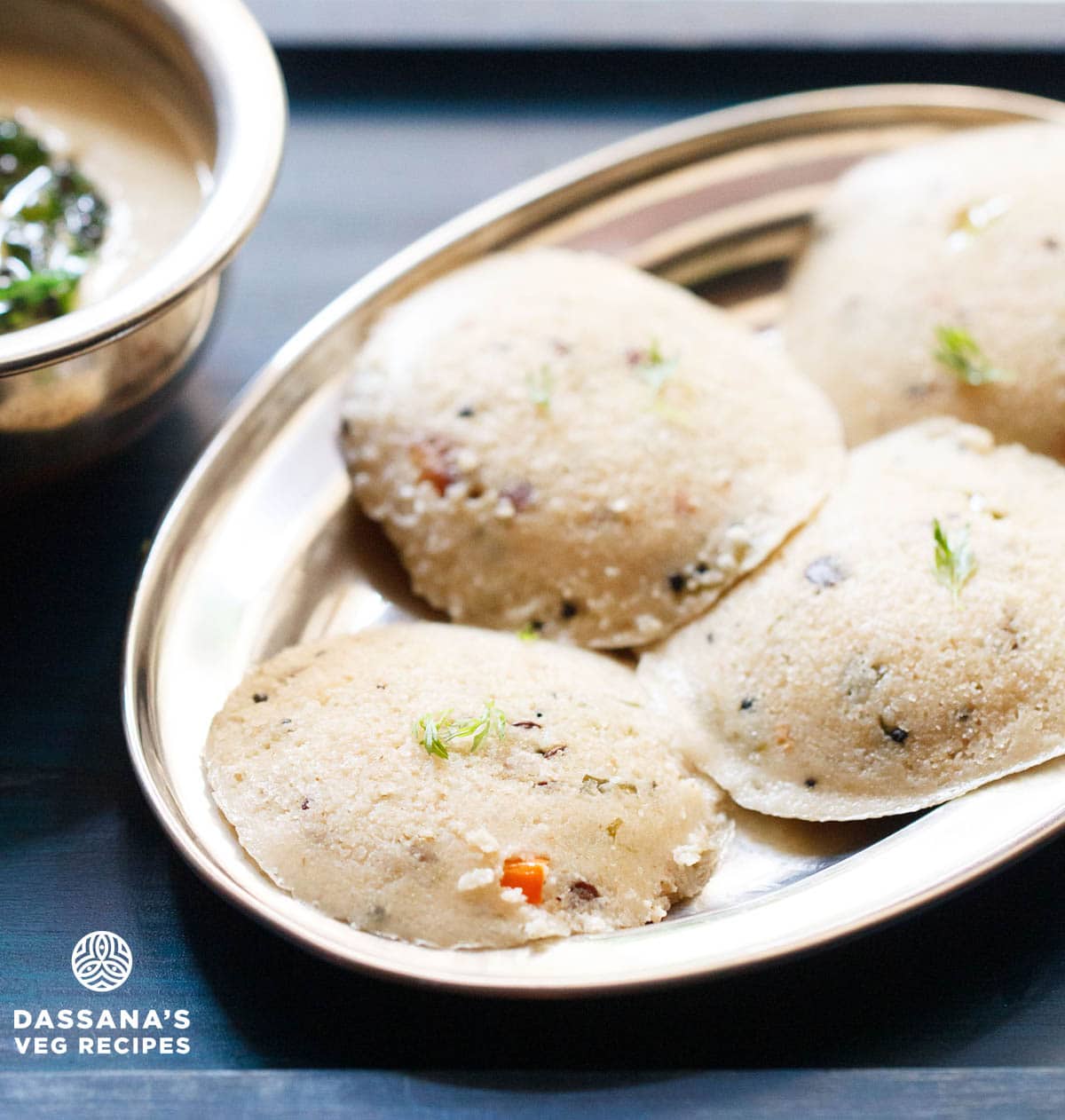 Four round oats idli arranged on silver plate with coconut chutney in a silver bowl.