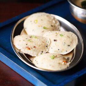 Four round oats idli arranged on silver plate on a dark blue wooden tray with coconut chutney in a silver bowl.