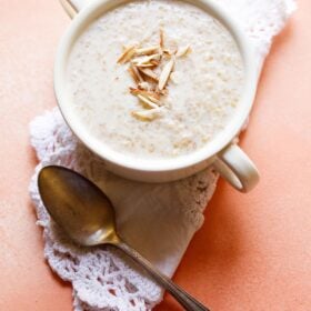 wheat porridge with slivered almonds in a white bowl with handles on a white folded doily with brass spoon below.
