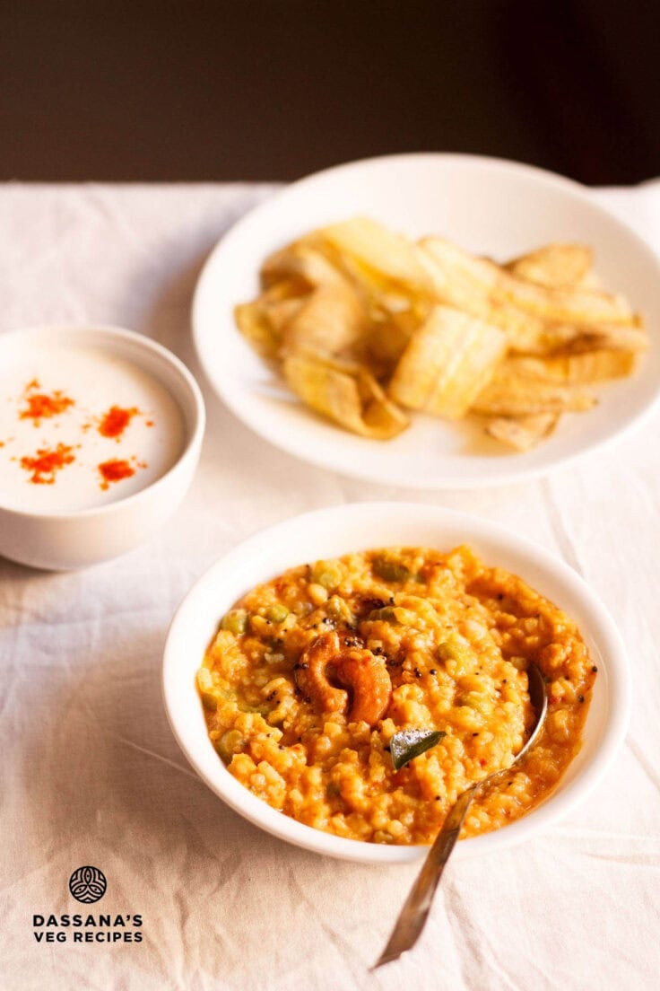 bisi bele bath served in a white bowl with a spoon inside, a bowl of banana chips kept on the top left side and a small bowl of raita kept on the top right side.
