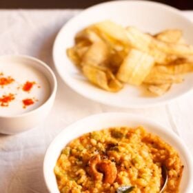 bisi bele bath served in a white bowl with a spoon inside, a bowl of banana chips kept on the top left side and a small bowl of raita kept on the top right side with text layovers.