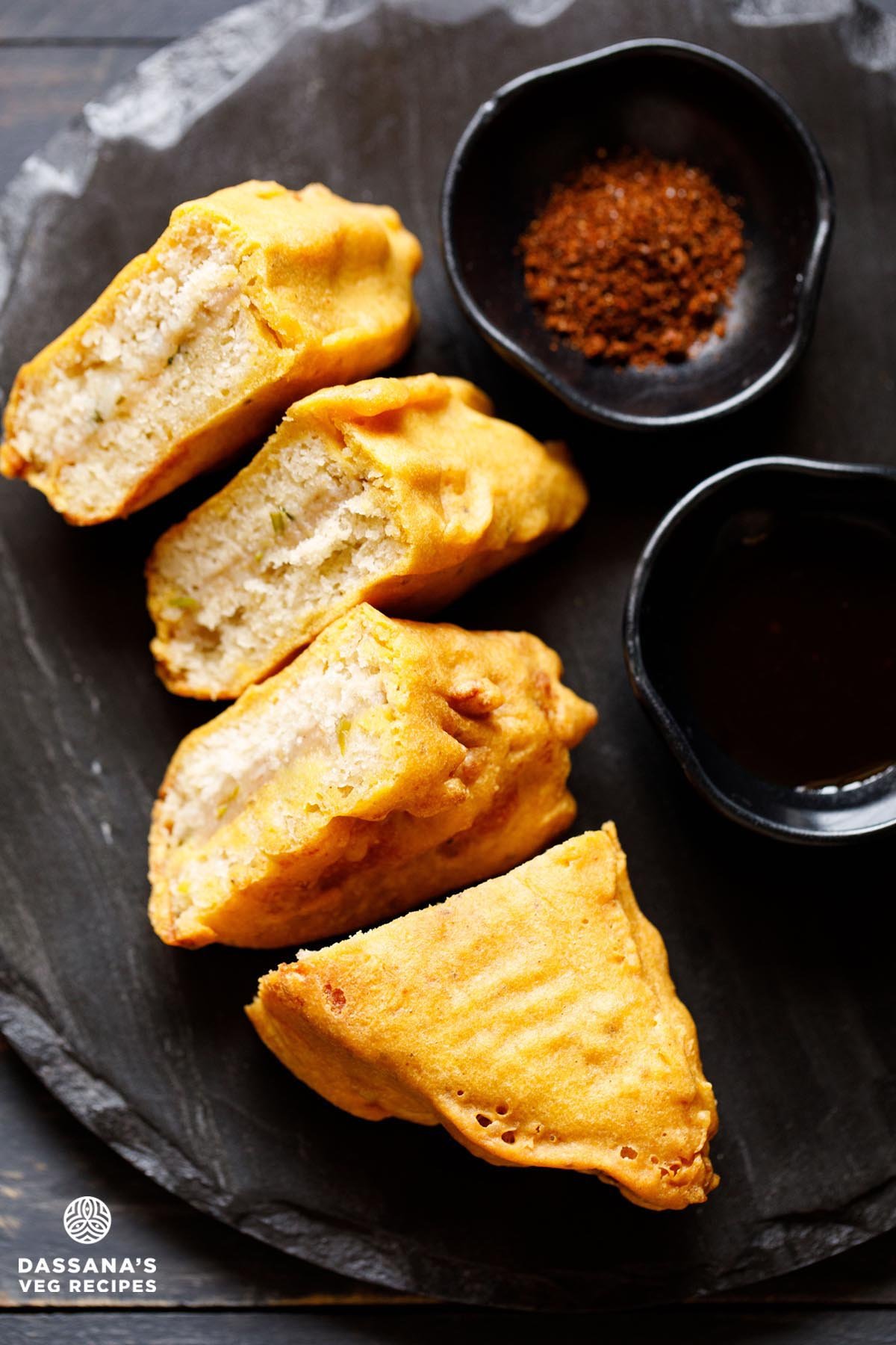 stuffed bread pakoda slices on a black slate board with two black small bowl of dry garlic chutney and tamarind chutney.