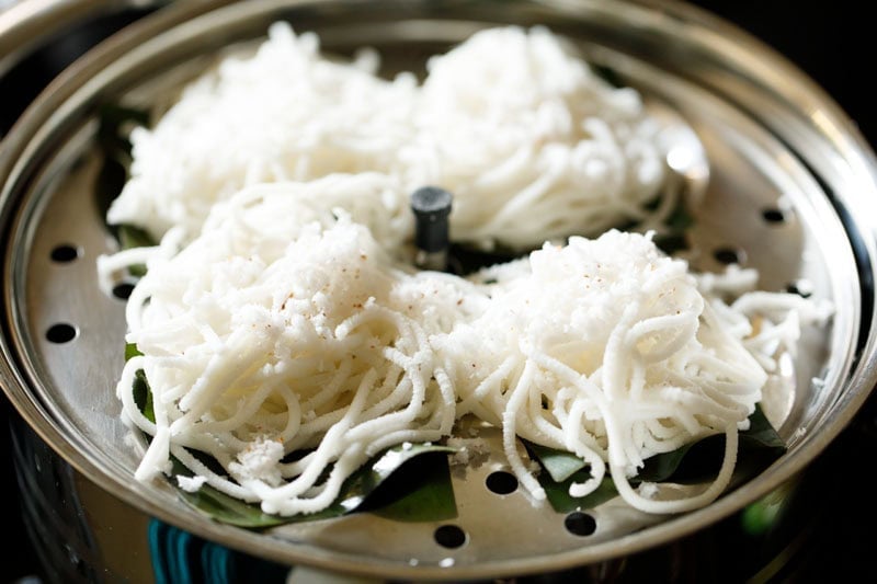 idiyappam rounds sprinkled with grated coconut on banana leaf squares on greased pan.