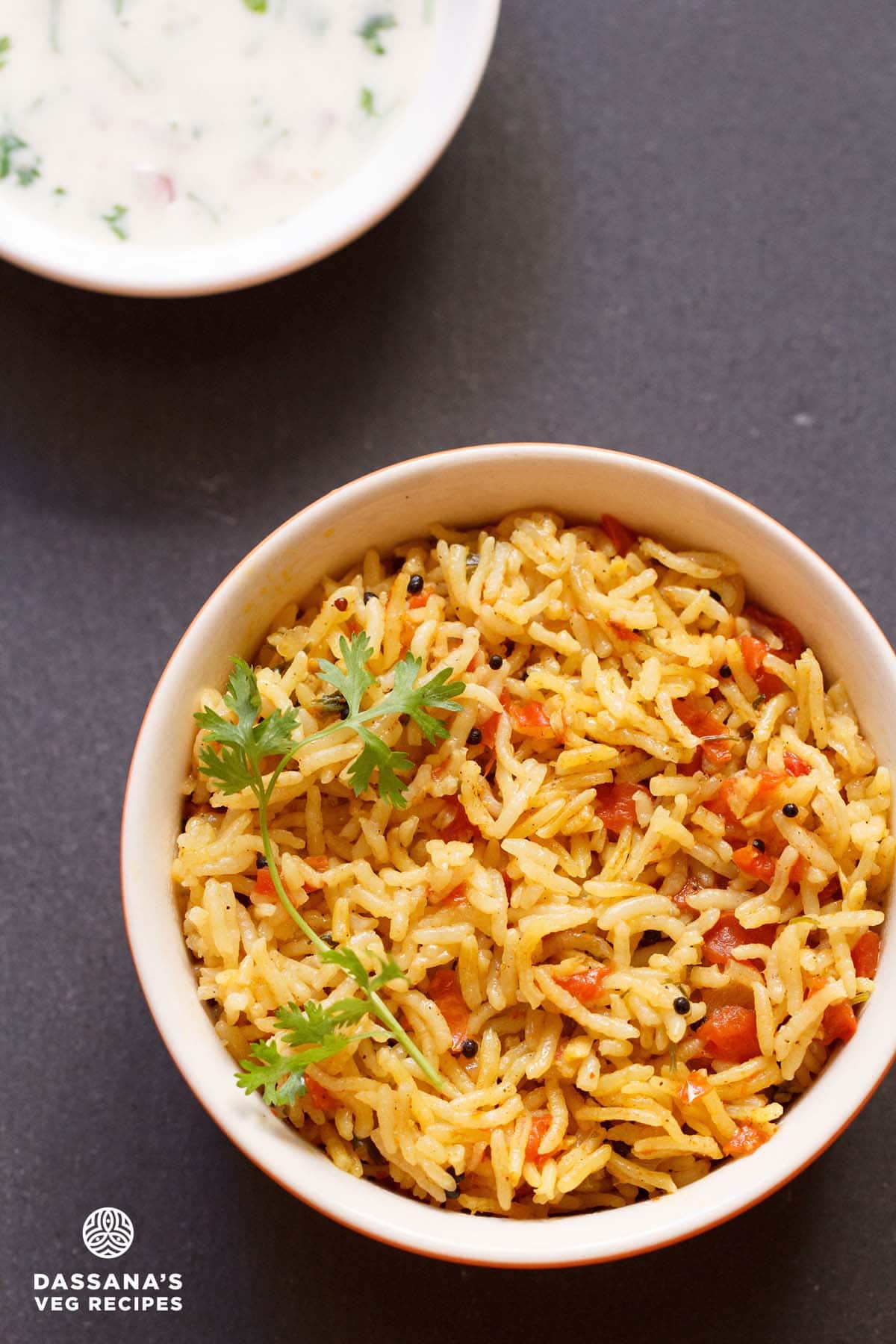 tomato rice with a sprig of cilantro served in an orange and red dotted bowl with a side yogurt raita on a dark black board