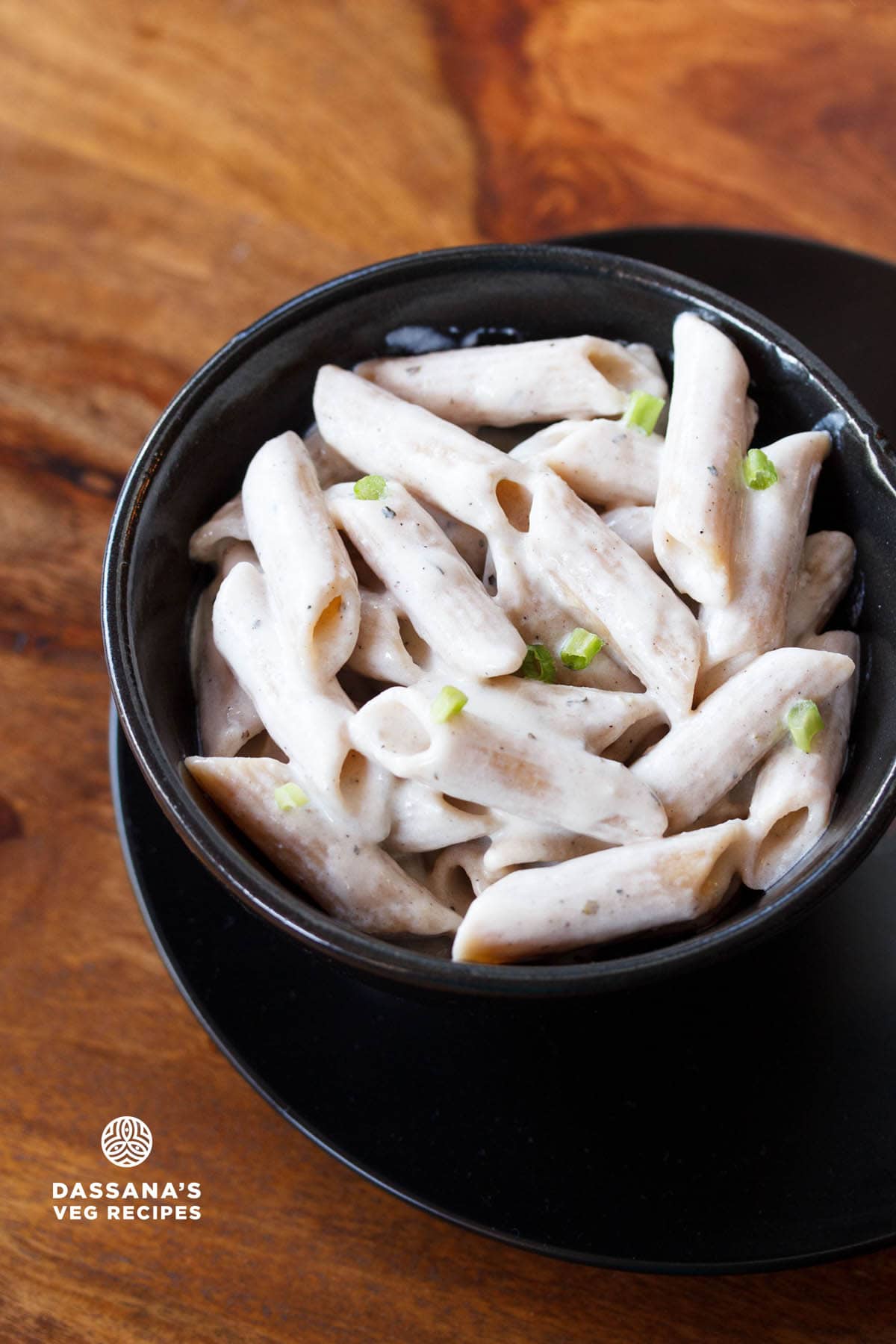 white pasta garnished with scallion greens served in a black glass bowl kept on top of black plate on a wooden board with text layovers. 