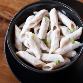 white pasta garnished with scallion greens served in a black bowl kept on top of black plate on a wooden board with text layovers.