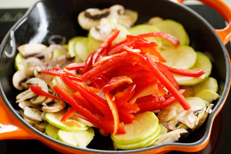 sliced red bell pepper on top of the sliced zucchini and mushrooms in skillet.