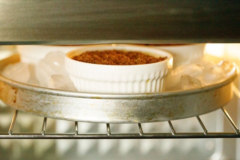 Ramekins of crème brûlée placed in a round baking pan filled with ice cubes under the oven broiler for caramelizing the sugar.