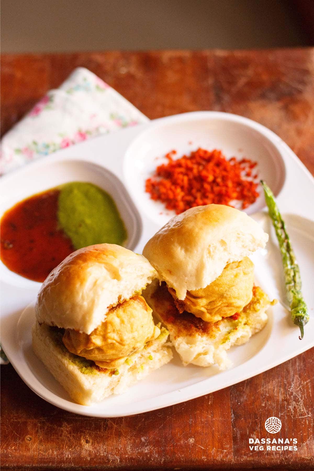 vada pav served with salted green chilies, dry garlic chutney and green chutney on a white plate with sections.