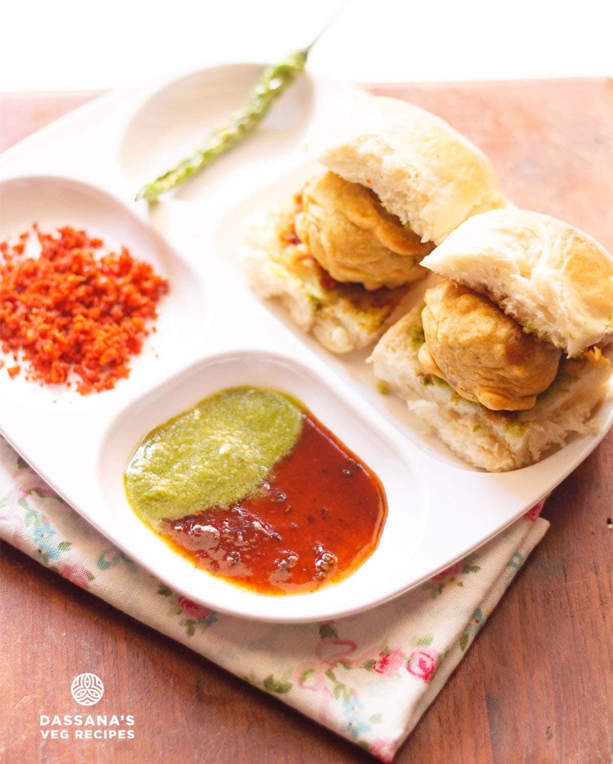 wada pav with green chilies, garlic chutney, green chutney and sweet chutney served in a white sectioned plate. 