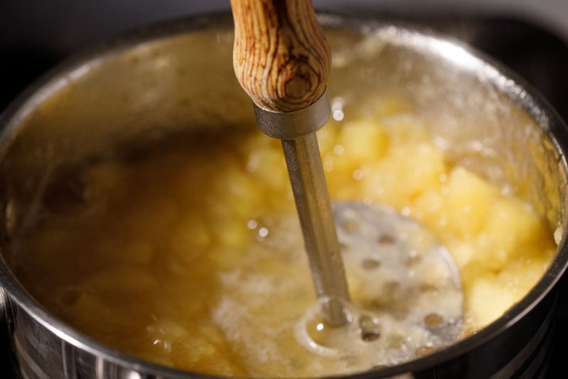 Cooked apples being mashed with a masher in pan.