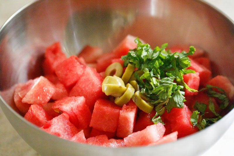 Sliced green olives and mint with watermelon cubes in mixing bowl.