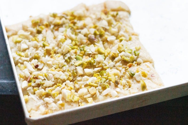 Malai barfi mixture spread on tray and topped with chopped nuts.