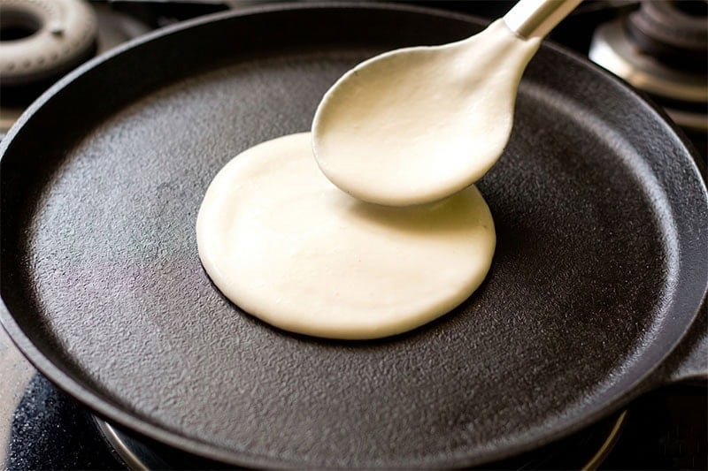 Fermented rice batter being spread with a spoon on a cast iron tawa.
